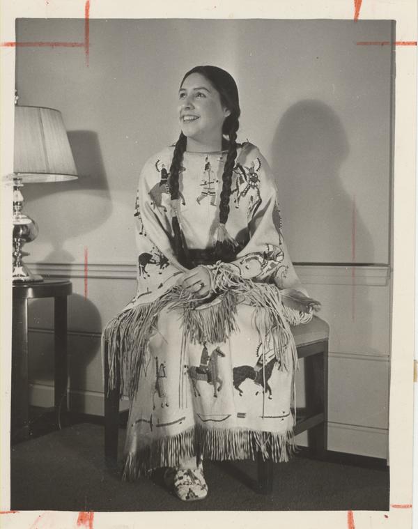 A black and white photo of Evelyn Yellow Robe, seated and wearing a traditional dress, decorated with images of people on horseback.  She is looking away from the camera, smiling.