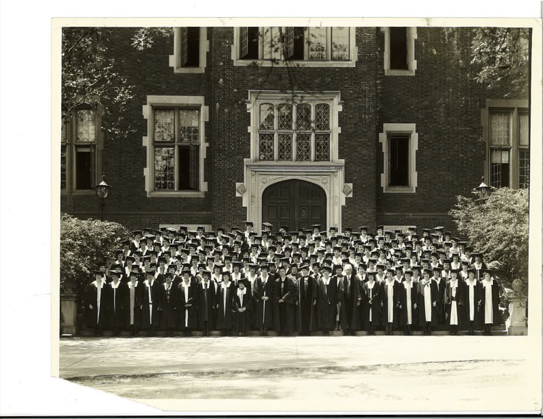 A group of students in caps and gowns stands before a brick building.