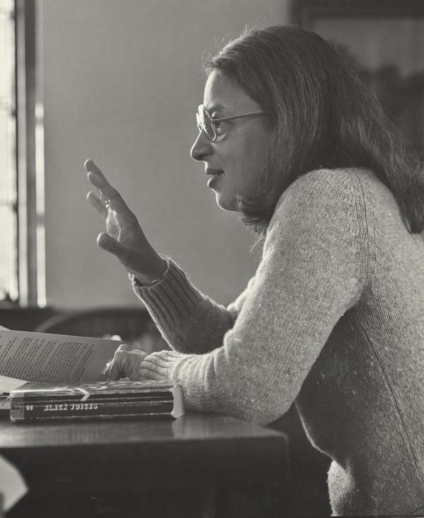 A Black woman with glasses and shoulder length hair sits at a table covered with books and papers.  
