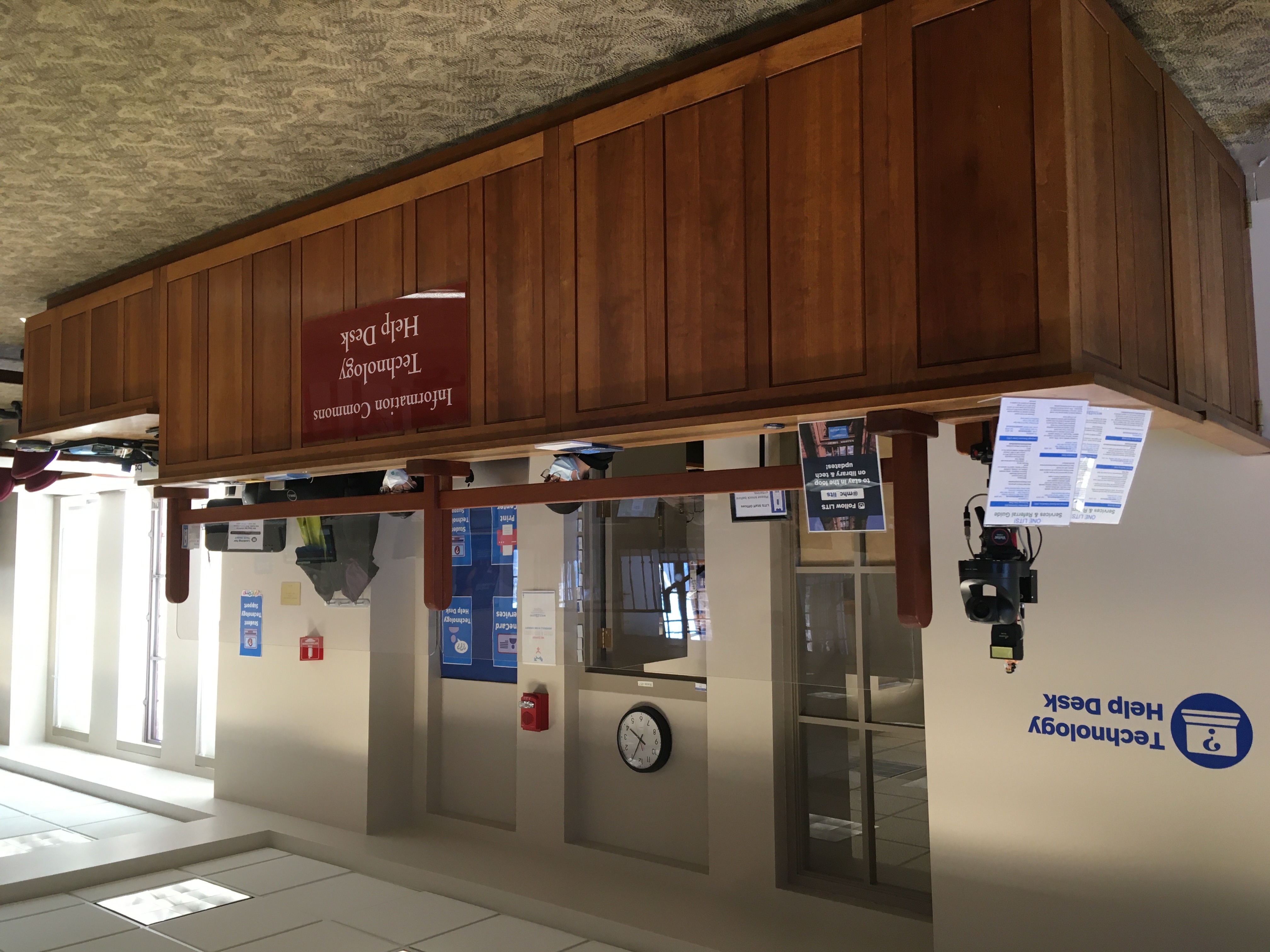 The Technology Help Desk on the fourth floor of the library. A large wooden desk with a plexiglass shield and student workers.