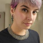 Yvaine is looking at the camera with a closed mouth smile. She has purple hair, she is wearing a black shirt, and is wearing a chainmail necklace.