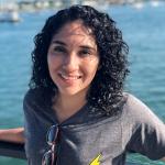 Maria is a woman with curly black hair wearing a gray shirt with a yellow lightning bolt on it. She is smiling and a body of water is behind her.