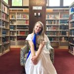 Aleah is a woman with blonde hair. She is wearing a white dress and a blue necktie. She is sitting in an armchair in the Williston Library's Reading Room.