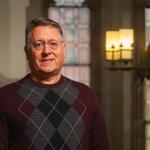 Ken is standing in front of a window. He has short gray hair, is wearing glasses, and is wearing a red patterned sweater.