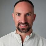 Brad is looking straight at the camera. He has blue eyes, brown hair, and a beard. He is smiling.