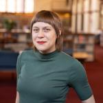 Abby is a woman with short brown hair and blue eyes. She is wearing a green short sleeved shirt and has red lipstick. She is smiling.