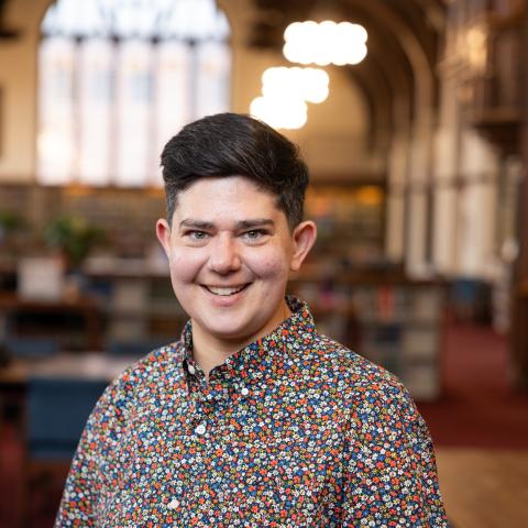 Caro has short, black hair and is wearing a button up shirt with small flowers on it. Caro is facing the camera and smiling.