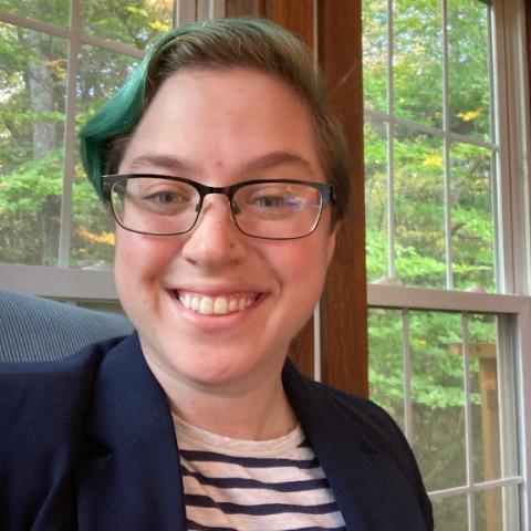 Skye is sitting in front of a window. They have short blue hair, are wearing glasses, and are smiling.